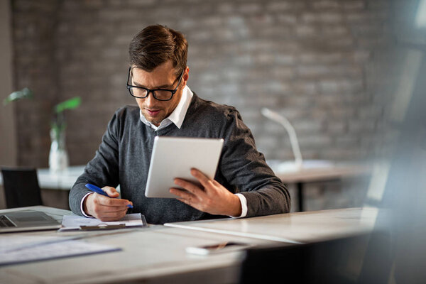 Male entrepreneur taking notes while going through paperwork and using touchpad at work. 