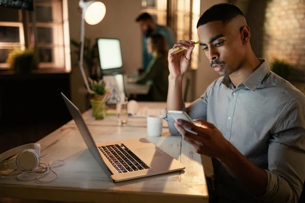 Young Black Businessman Using Mobile Phone While Working Computer Evening — Zdjęcie stockowe