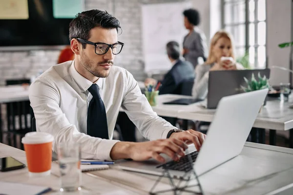 Young entrepreneur typing an e-mail while using computer in the office. There are people in the background.