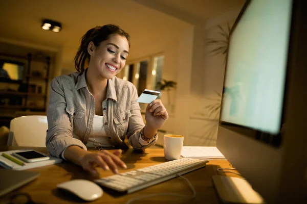 Young smiling woman using computer and credit card while buying on the internet at night at home.