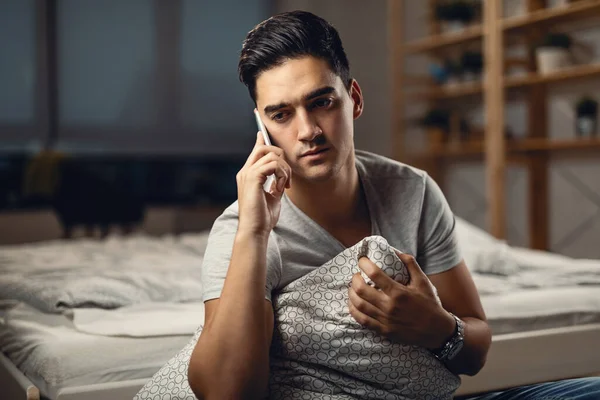 Sad man communicating over cell phone while sitting in the bedroom and thinking of something.