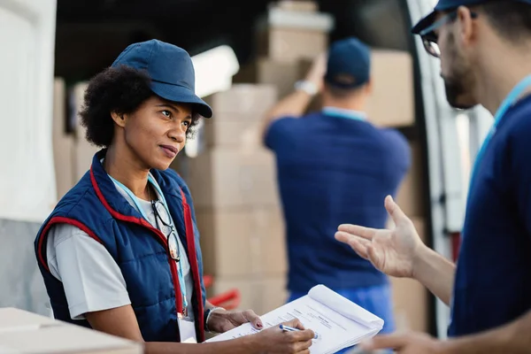 Two deliverers communicating and going through paperwork while their colleagues is loading packages in a van. Focus is on African American woman.