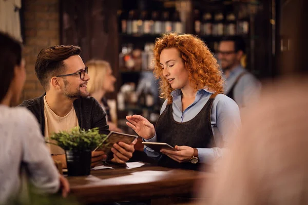 Redhead waitress communicating with a guest while taking his order and using touchpad in a bar.