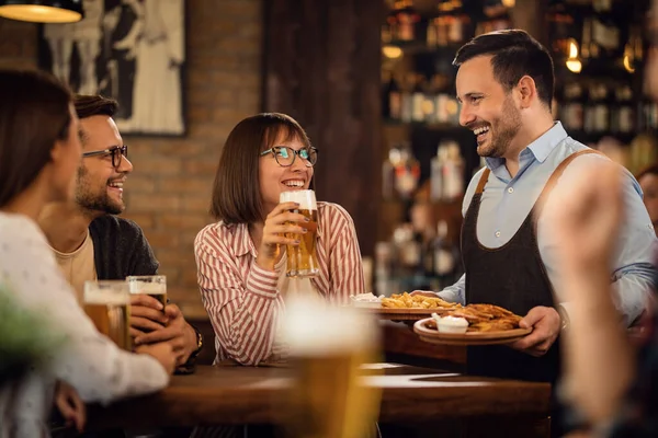 Happy waiter talking with guests who are drinking beer while brining food at their table.