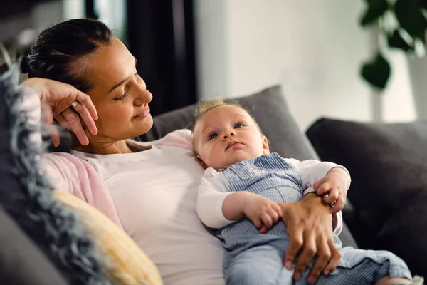 Smiling mother holding her baby boy and looking at him with affection while relaxing on sofa at home. 