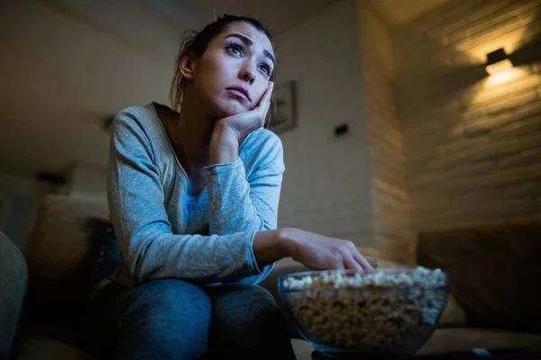 Low angle view of sad woman watching TV and eating popcorn in the evening at home.