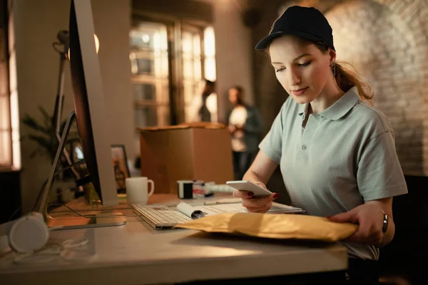 Young deliverer scanning bar code on package with mobile phone while working in the office.