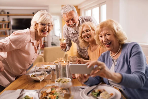 Group of happy seniors using smart phone and taking selfie at dining table at home.