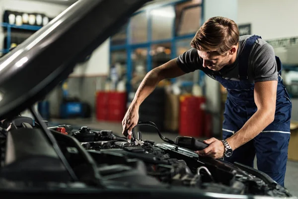 Auto mechanic using diagnostic tool and attaching jumper cables on car engine in a repair shop. 