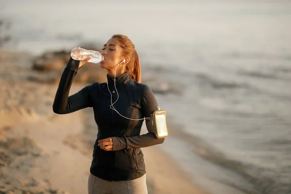 Young sportswoman drinking water from a bottle while taking a break from morning run at the beach,