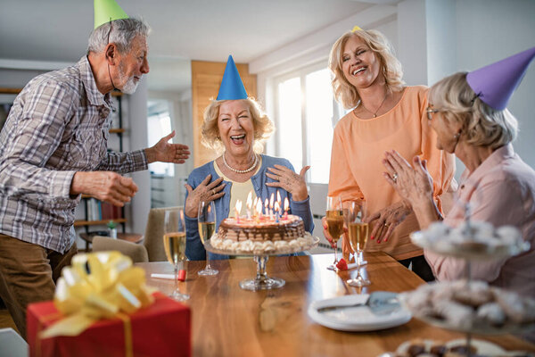 Веселая пожилая женщина празднует день рождения с друзьями и весело проводит время на домашней вечеринке. 