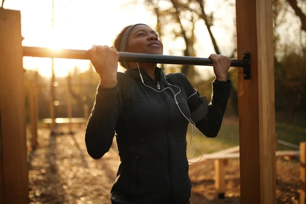 American American woman practicing chin-ups while exercising at outdoor gym at sunset.