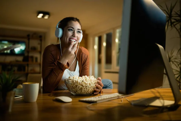 Young smiling woman relaxing in the evening at home while eating popcorn and watching something on a computer. 