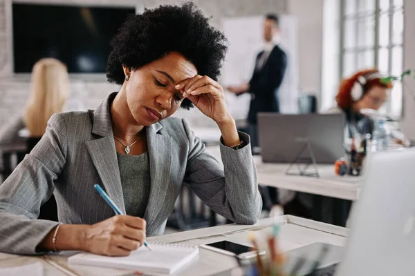 African America female entrepreneur making a plan and writing notes while working at her desk in the office. There are people in the background.