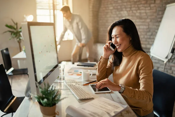 Happy Asian entrepreneur working on computer while talking on the phone at her office desk.