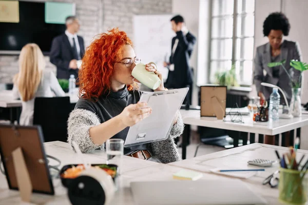 Female entrepreneur reading business reports and drinking coffee while sitting at her desk in the office. There are people in the background.