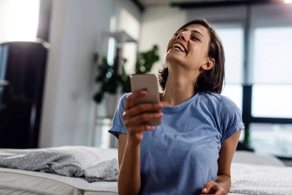 Young woman laughing while reading text message on mobile phone in the bedroom.