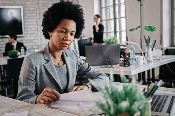 Black female entrepreneur preparing for upcoming meeting while going through her notes in the office. There are people in the background.