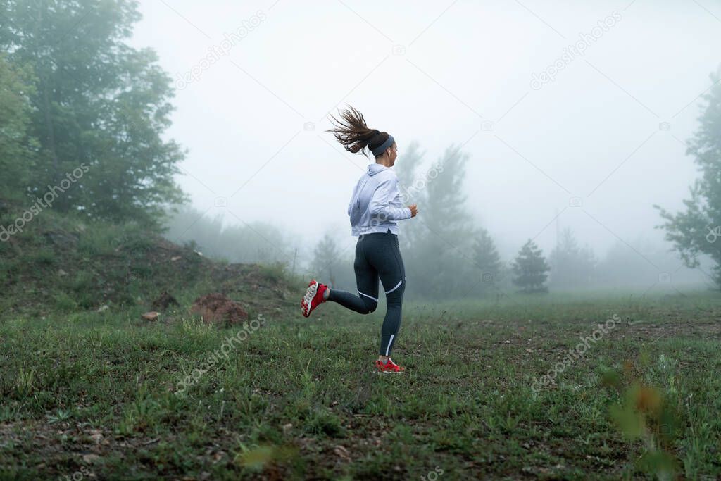 Athletic woman jogging in nature on foggy morning and listening music over earphones. Copy space.