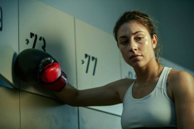 Sweaty athletic woman thinking of something in gym's dressing room after boxing training. 