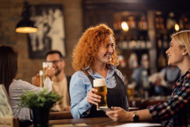 Young happy waitress serving beer and communicating with female guest in a bar. 