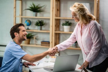 Happy doctor shaking hands with senior woman who came to medical appointment. 