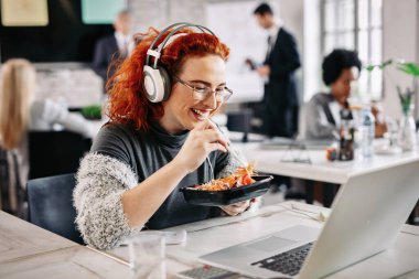 Cheerful freelancer using computer and eating salad on a lunch break in the office. 