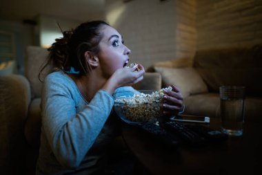 Young woman feeling hungry and eating popcorn from a bowl while watching movie at night at home. 