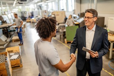 Happy engineer visiting steel factory and shaking hands with African American worker while using touchpad. 