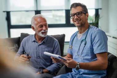 Happy male doctor visiting senior patients at home and going through medical documents. 
