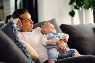 Pensive mother and her baby boy sitting on sofa in the living room and looking away.