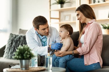 Smiling mother holding her small son while doctor is examining him with stethoscope during medical appointment. 