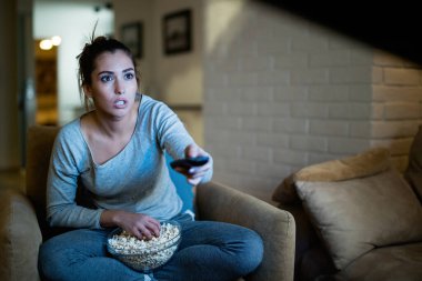 Young woman in disbelief changing channels with remote control while watching TV and eating popcorn at night. 