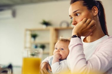 Young woman suffering from postnatal depression and thinking of something while being with her baby at home.