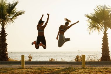 Rear view of playful sportswomen having fun while jumping with their arms raised at sunset. 