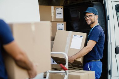 Young happy manual worker carrying cardboard boxes in delivery van while communicating with his colleagues. 