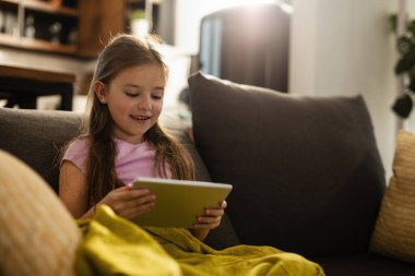 Cute small girl surfing the Internet while sitting on the sofa at home. 