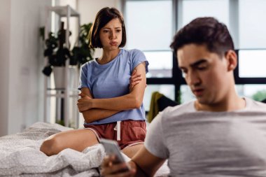 Young displeased woman sitting on bed with arms crossed and looking at her boyfriend after the argument. 