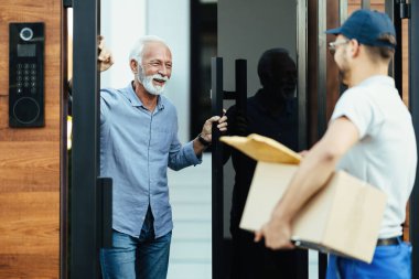 Happy mature man standing at the gate and talking with courier who is delivering him a package. 