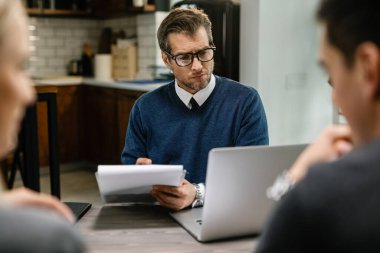Serious financial advisor writing notes and using laptop while having a meeting with a couple at their home. 