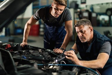 Two mechanics cooperating while working on air conditioning system of a car in auto repair shop. 