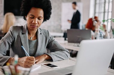 African American businesswoman working in the office an taking notes while preparing for upcoming meeting. There are people in the background.