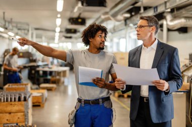 Company manager holding project plans while talking to African American manual worker who is pointing at something in the factory. 