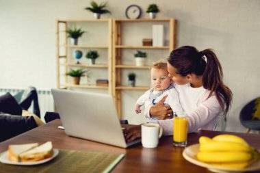 Young mother holding her baby boy while working on laptop at home. 