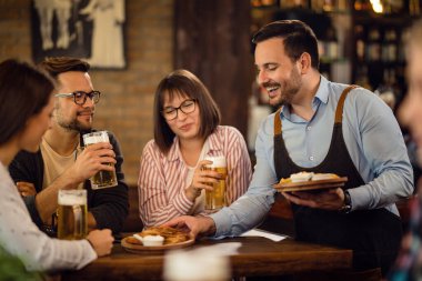 Group of happy friends enjoying in a glass of beer while waiter is serving food at their table in a pub. 