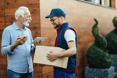 Happy mature man talking to young deliverer while signing on digital tablet for home delivery. 