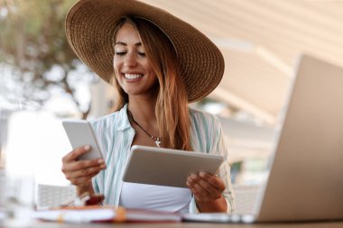 Young smiling woman using digital tablet while reading text message on mobile phone in a cafe.