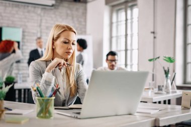 Mid adult businesswoman using laptop and reading an e-mail at work. There are people in the background. 