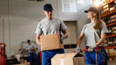 Two happy coworkers communicating while working with packages in factory warehouse. Focus is on man. 