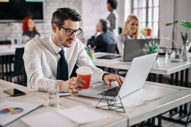 Young businessman sitting in the office and working on laptop while drinking coffee. There are people in the background. 